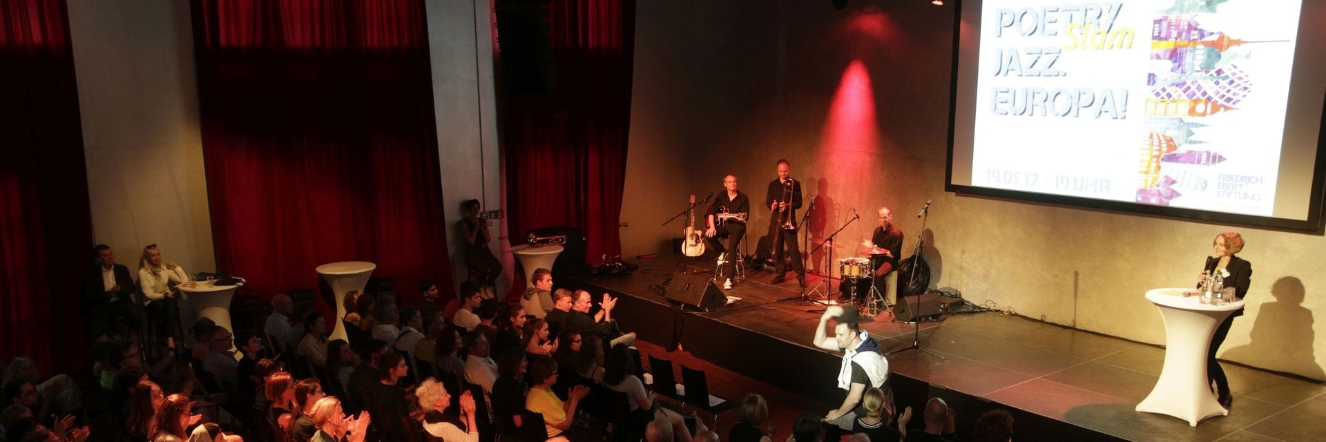Ein Veranstaltungssaal mit einer Bühne, auf der eine Musikgruppe mit Instrumenten auftritt. Rechts auf der Bühne steht eine Person an einem Rednerpult. Im Publikum sitzen zahlreiche Menschen, die applaudieren. Im Hintergrund hängen rote Vorhänge, und auf einer Leinwand ist der Titel „Poetry Slam Jazz Europa!“ mit weiteren Informationen zu sehen.