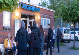 Teilehmende stehen und reden vor dem Veranstaltungsort mit dem Schriftzug "Türkiyemspor" im Hintergrund