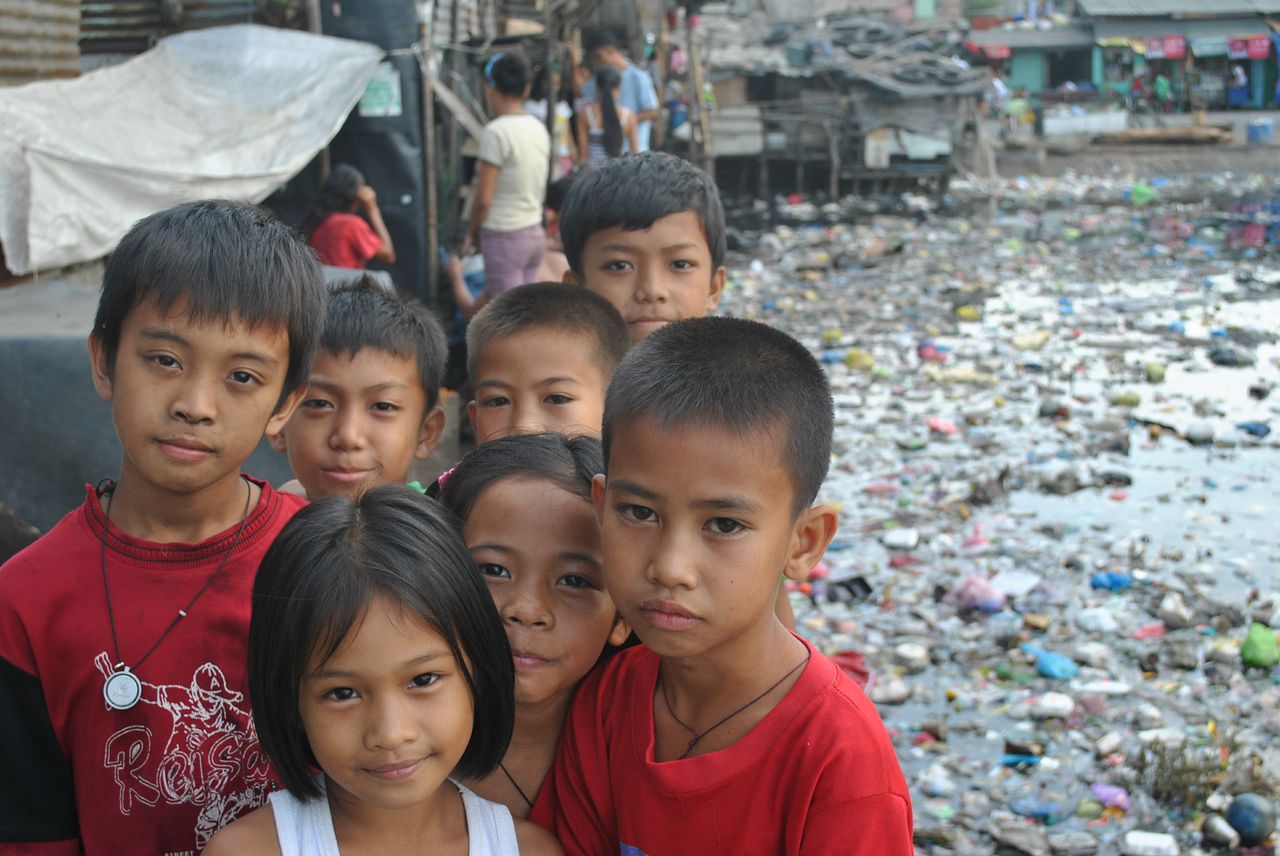 Philippinische Kinder schauen in die Kamera, im Hintergrund ärmliche Gegend und vermülltes Wasser