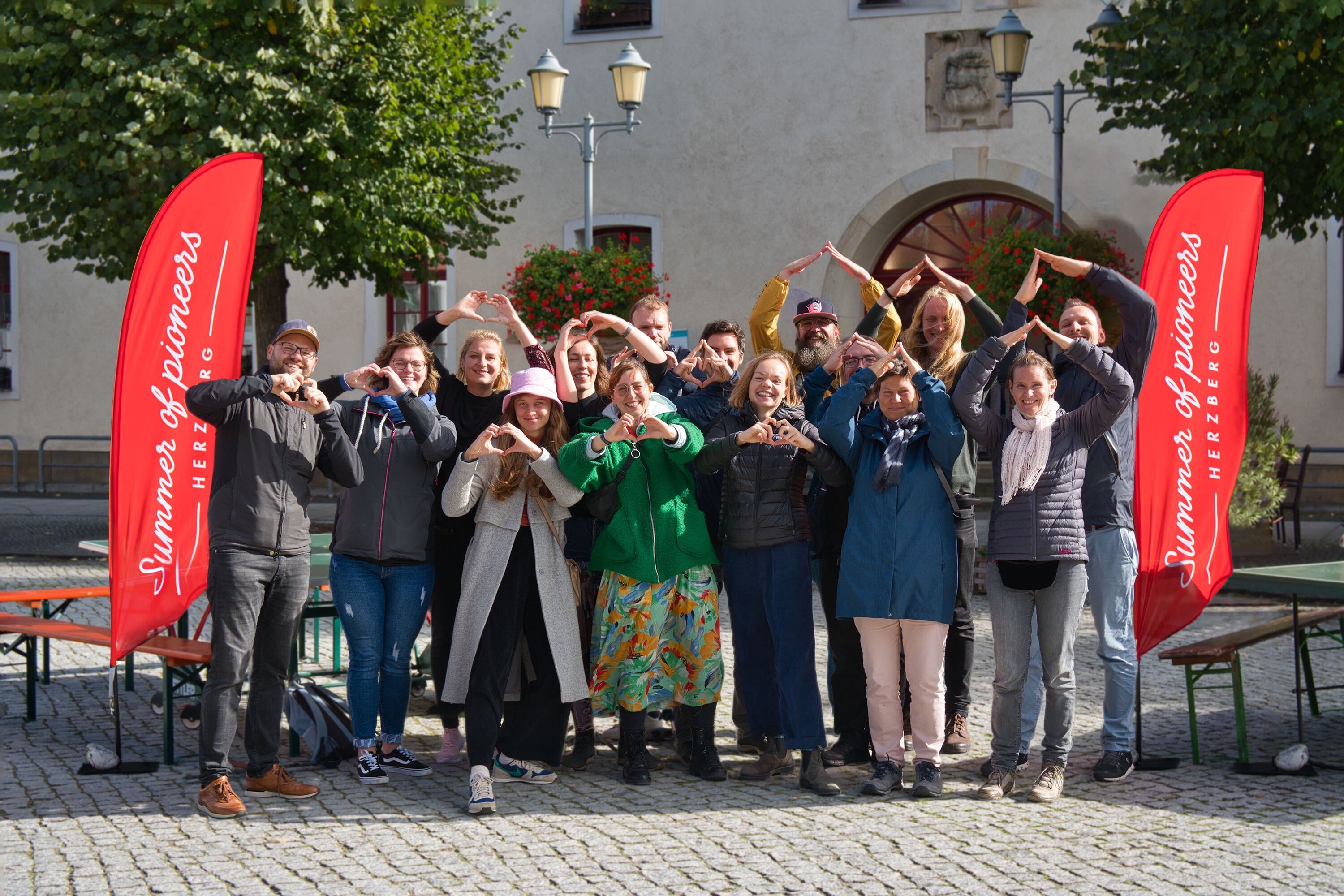 Eine Gruppe von 15 Frauen und Männern steht vor einem Gebäude zwischen zwei roten Fahnen mit der Aufschrift "Summer og Pioneers". Mehrere Personen formen ihre beiden Hände zu einem Herz.