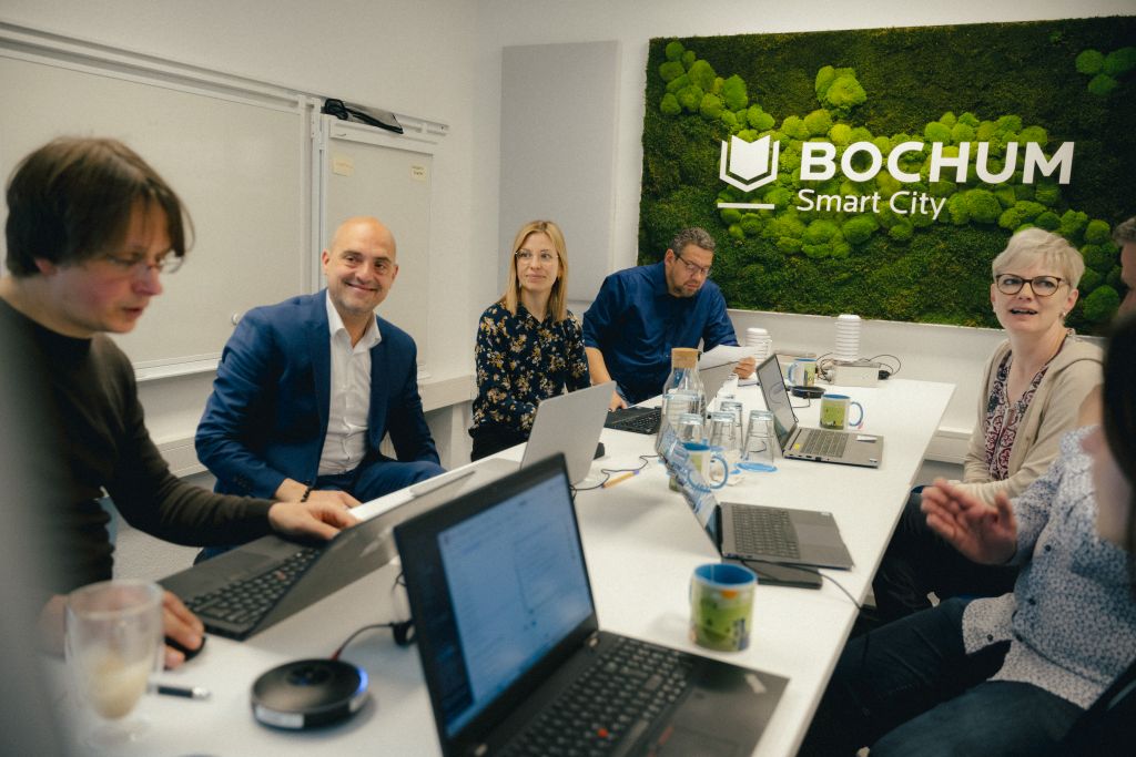 Fünf Personen sitzen mit Laptops um einen rechteckigen Tisch, an der Wand das Logo von Bochum Smart City