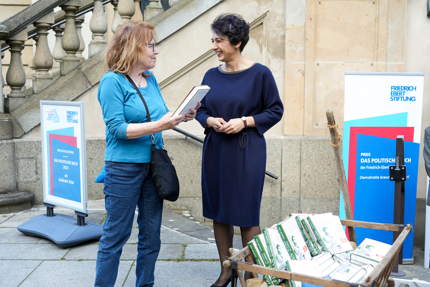 Die Preisträgerin des Politischen Buches 2023 Golineh Atai im Austausch mit einer Passantin, die ein Buch in der hand hält. Die beiden stehen vor einer Steintreppe, neben sich ein Holzkarren voller Bücher und ein Roll-Up der Friedrich-Ebert-Stiftung.