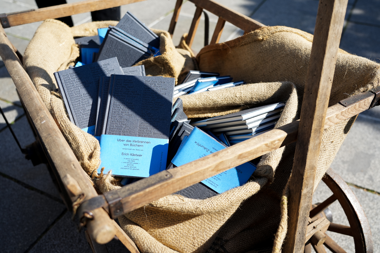 Ein Holzkarren voller Bücher, die auf Sackleinen liegen. Im Vordergrund ist Erich Kästners "Über das Verbrennen von Büchern" zu sehen.