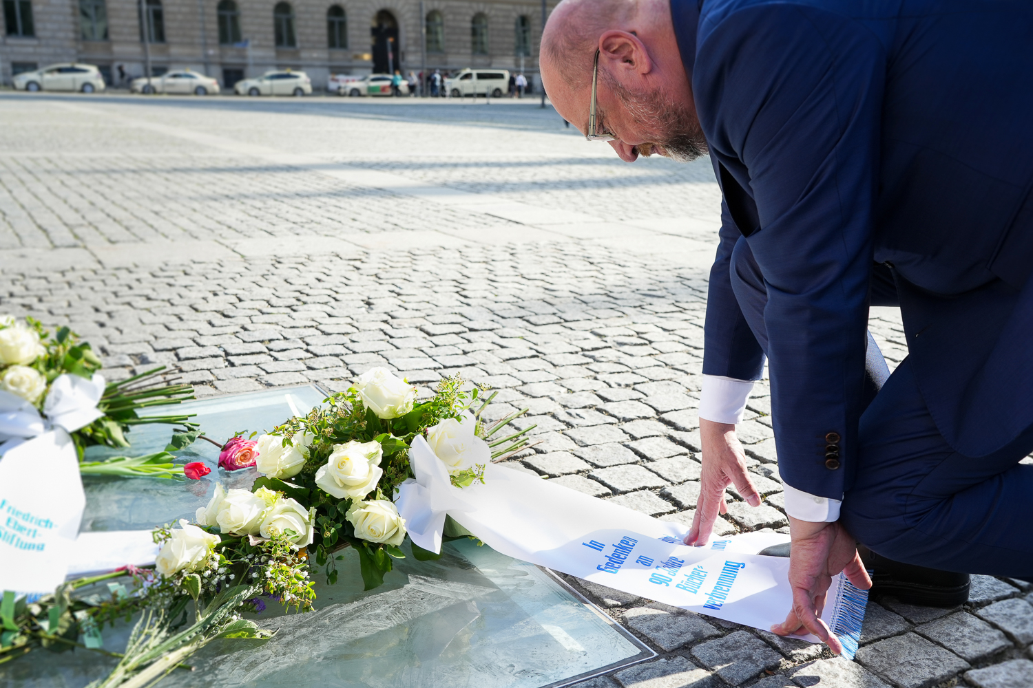 Nahaufnahme von Martin Schulz, der auf einem Platz vor einem Kranzgebinde mit der Aufschrift "In Gedenken an 90 Jahre Bücherverbrennung" kniet