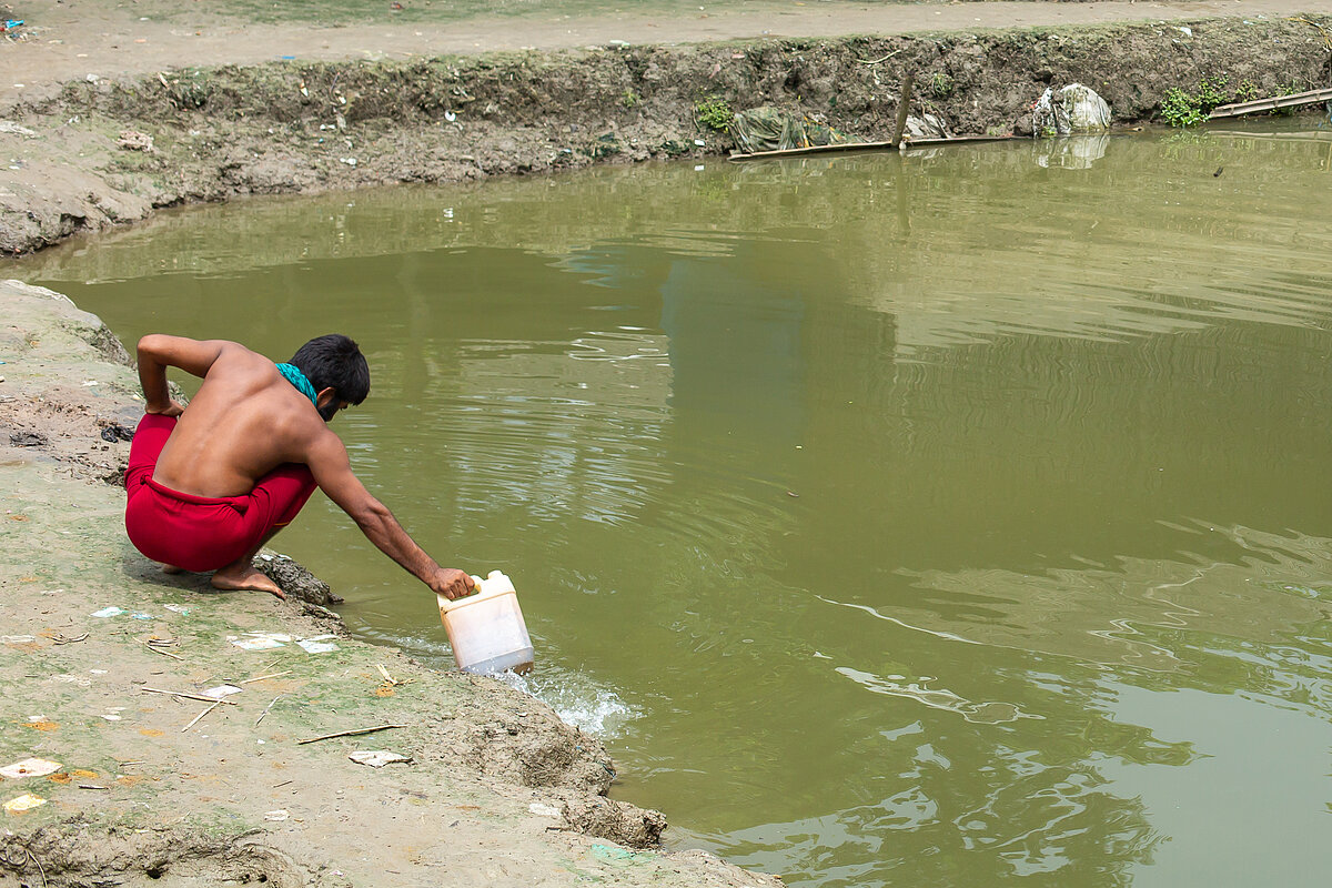 Ein Mann aus Bangladesch schöpft Wasser zum Trinken aus einem grünen Teich. 