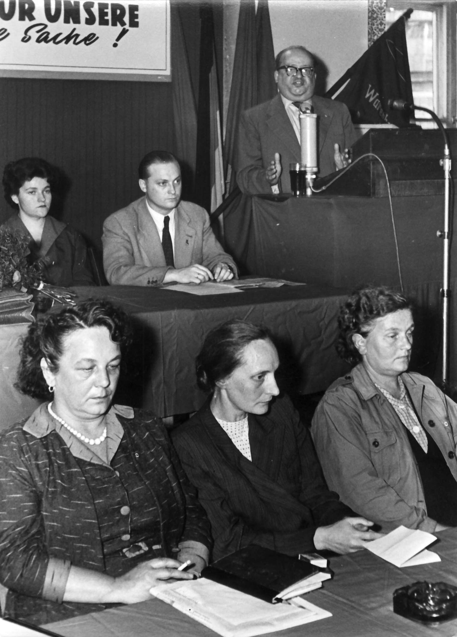 Friedrich Ebert im Hintergrund an einem Rednerpult, im Vordergrund in zwei Reihen Zuhörer:innen vermutlich auf einem Podium