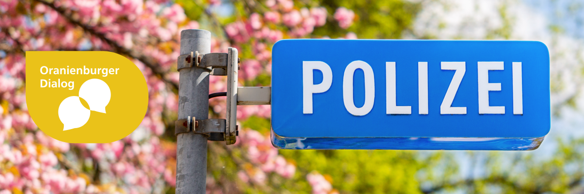 Abbildung: Metallstange mit einem daran befestigten Hinweisschild in blau mit weißem Schriftzug "POLIZEI". Im Hintergrund sind unscharf rosa blühende Bäume erkennbar. Der Himmel ist blau und leicht bewölkt