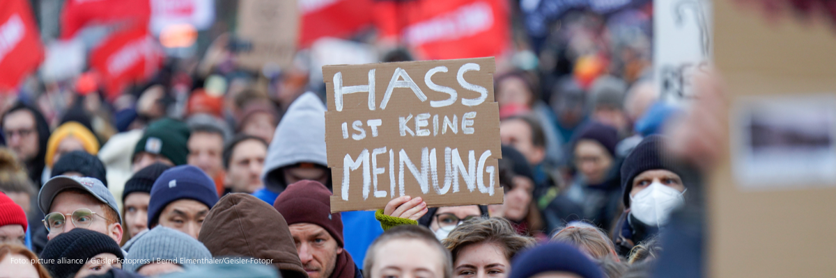 eine große Menschenmenge auf einer Demonstration gegen Rechts. In der Mitte hält jemand ein Pappschild mit der Aufschrift "Hass ist keine Meinung". Unscharf im Hintergrund sind viele rote Fahnen zu sehen. 