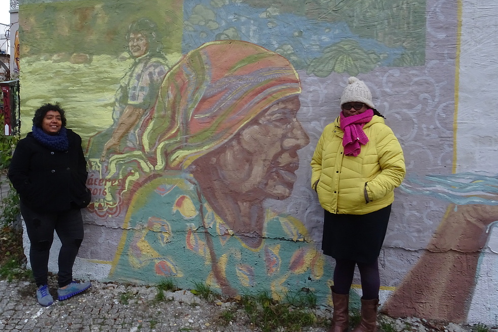 Miriam Miranda vor einem Graffiti von Berta Cáceres. Graffiti stellt eine ältere Frau mit Kopftuch dar