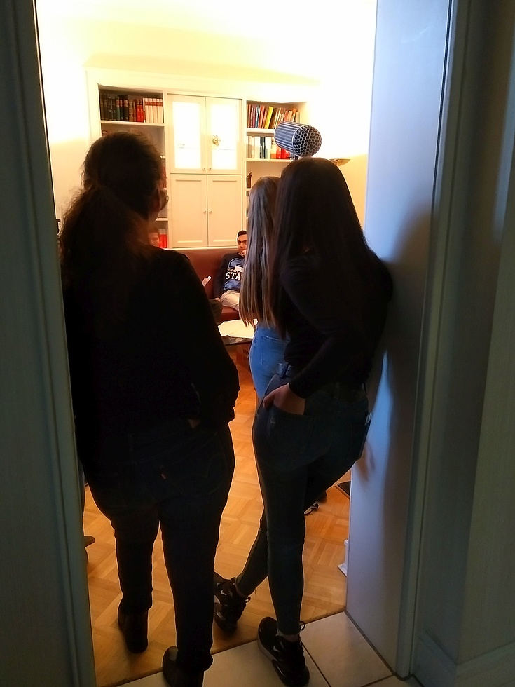 Zwei Mädchen stehen mit einem Mikrofon und einer Kamera im Türrahmen und nehmen das Gespräch zweier Männer auf dem Sofa auf.