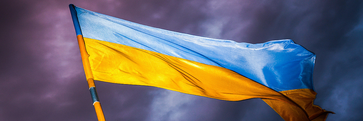Wehende Ukraineflagge am Fahnenmast vor einem dunklen Hintergrund.