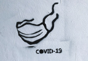 schwarzes Grafitti auf einer weißen Wand. Ein angedeutetes Gesicht mit einer FFP2 Maske und dem Schriftzug "COVID-19"