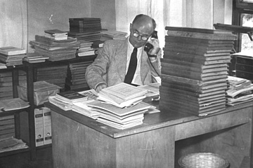Rudolf Rothe, Herr mit Brille, telefoniert mit einem Kabeltelefon, hinter einem Schreibtisch sitzend, umgeben von hohen Bücherstapeln