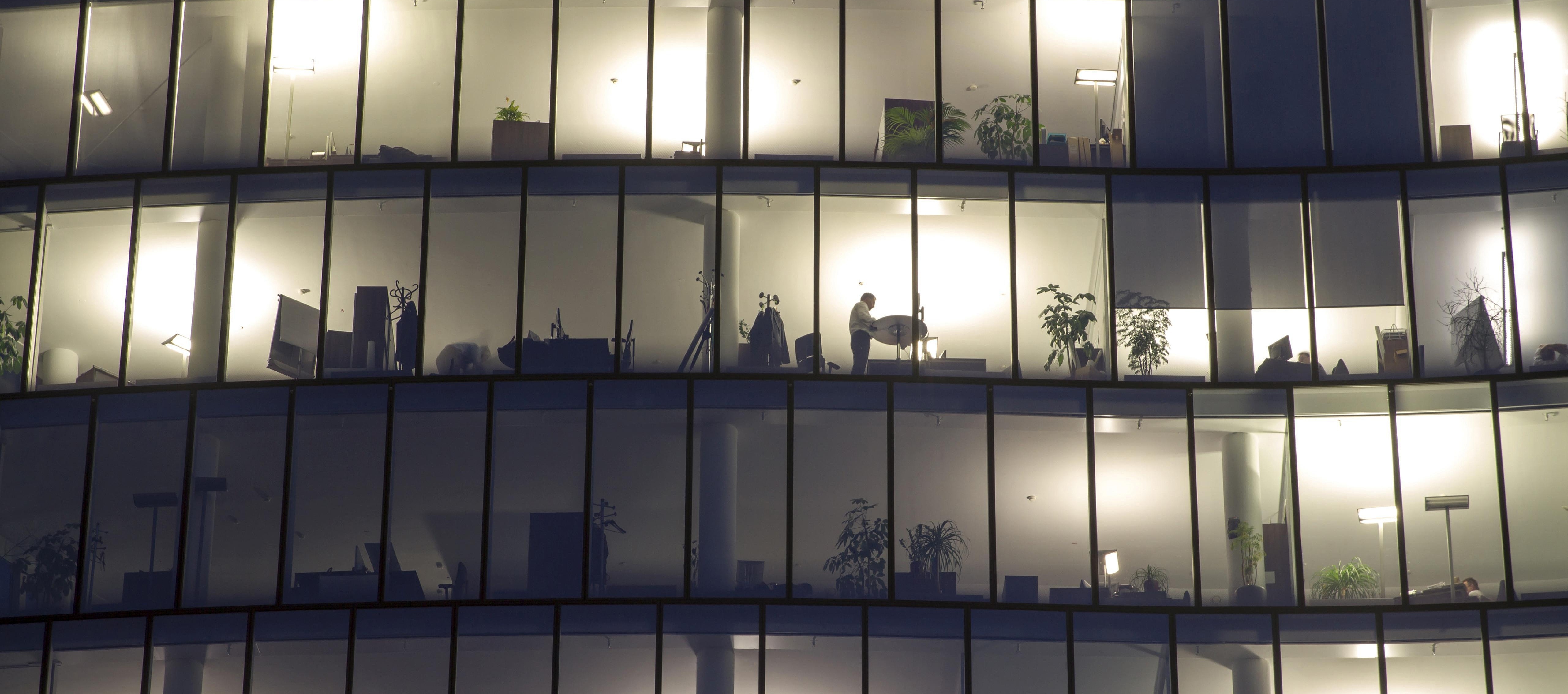 Blick auf eine mehrstöckige Fensterfront, hinter der vereinzelt Personen vor oder an ihren Arbeitsplätzen zu sehen sind.