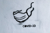 schwarzes Grafitti auf einer weißen Wand. Ein angedeutetes Gesicht mit einer FFP2 Maske und dem Schriftzug "COVID-19"