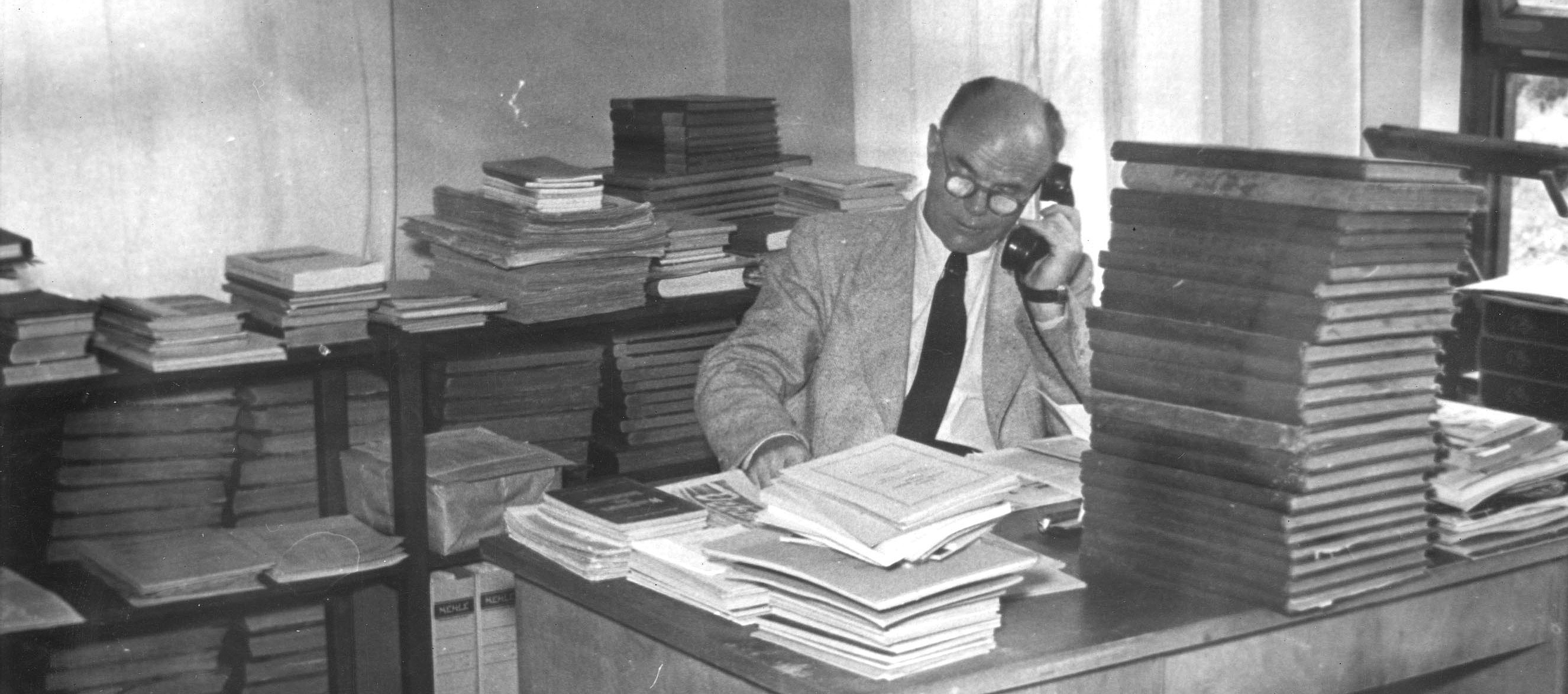 Rudolf Rothe, Herr mit Brille, telefoniert mit einem Kabeltelefon, hinter einem Schreibtisch sitzend, umgeben von hohen Bücherstapeln