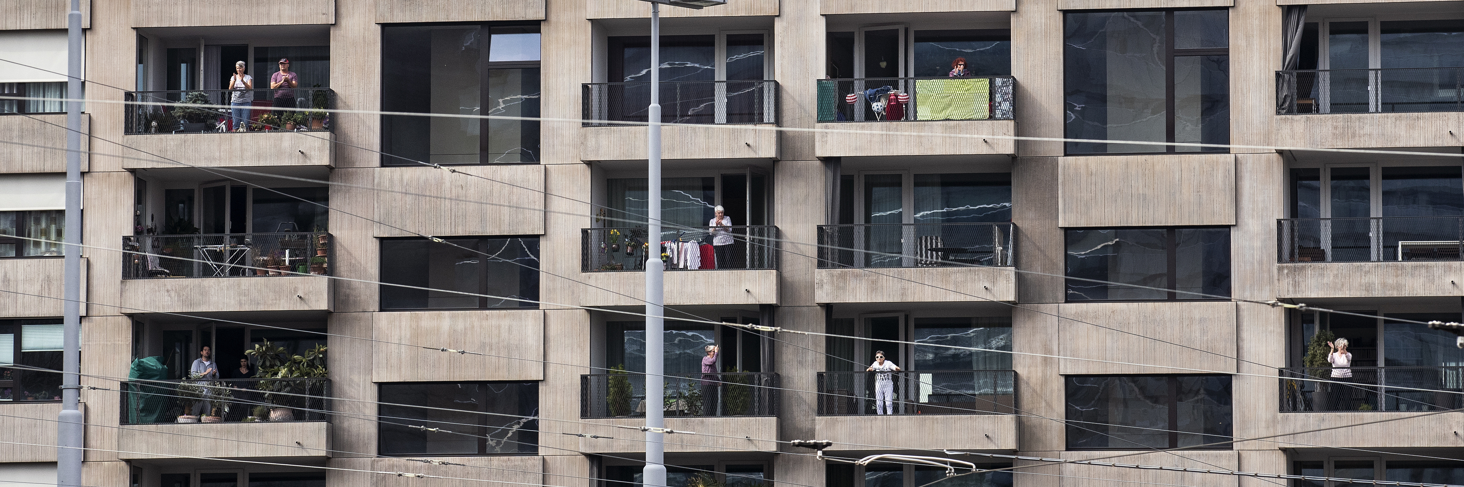 Mehrstöckiges Wohnhaus mit grauer Fassade. Einzelne Bewohnerinnen und Bewohner stehen auf Balkonen neben Wäscheständern und Topfpflanzen und blicken auf die Straße.