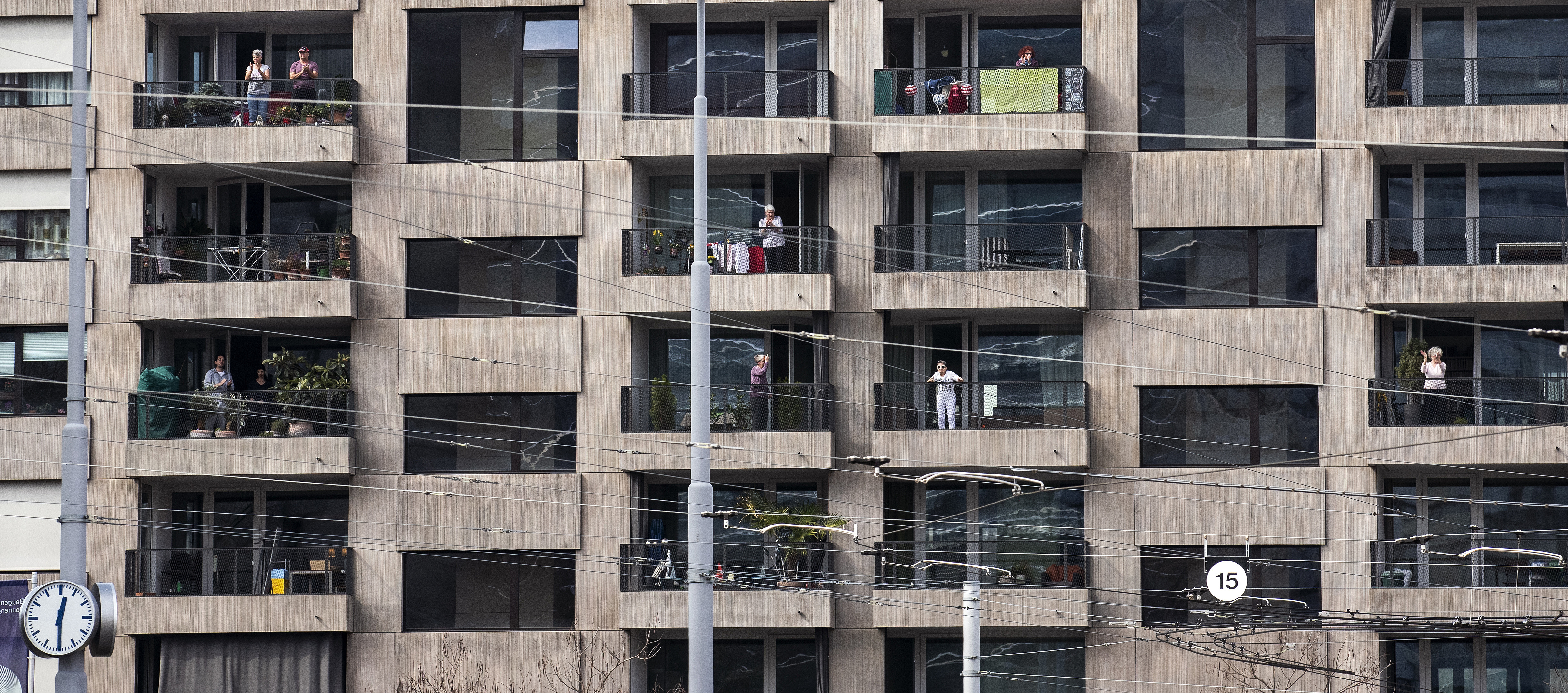 Mehrstöckiges Wohnhaus mit grauer Fassade. Einzelne Bewohnerinnen und Bewohner stehen auf Balkonen neben Wäscheständern und Topfpflanzen und blicken auf die Straße.