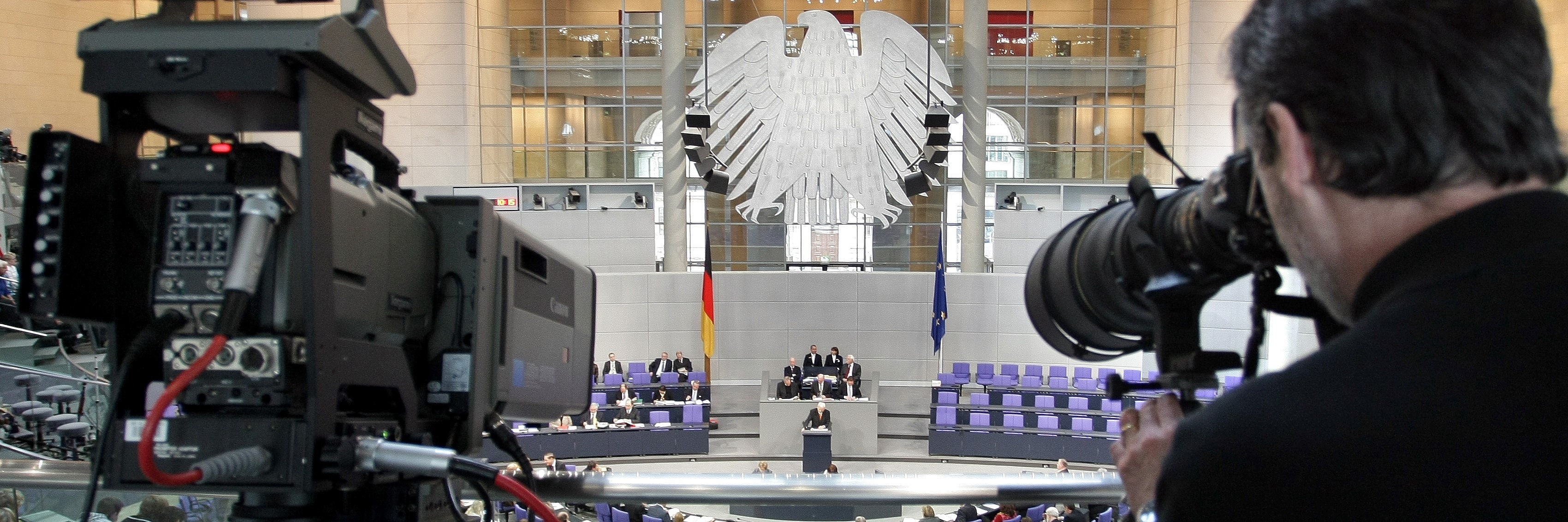 Von der Pressetribüne des Plenarsaals im Deutschen Bundestag fotografiert ein Journalist in Richtung des Redner_innenpultes. Unten im Saal sind die Abgeordneten, vor allem aber das Podium der Sitzungsleitung und die Sitzreihen der Bundesregierung zu sehen. 