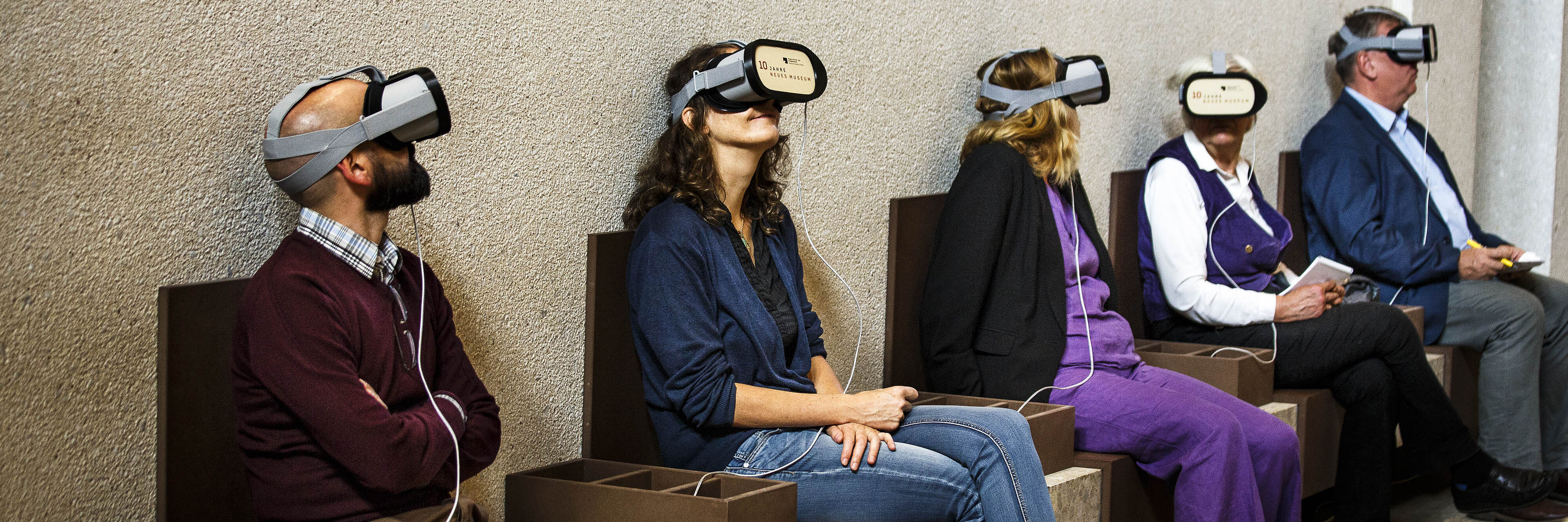 Fünf Personen unterschiedlichen Geschlechts und Alters mit Virtual-Reality-Headsets sitzen nebeneinander auf einer Bank in einem Raum