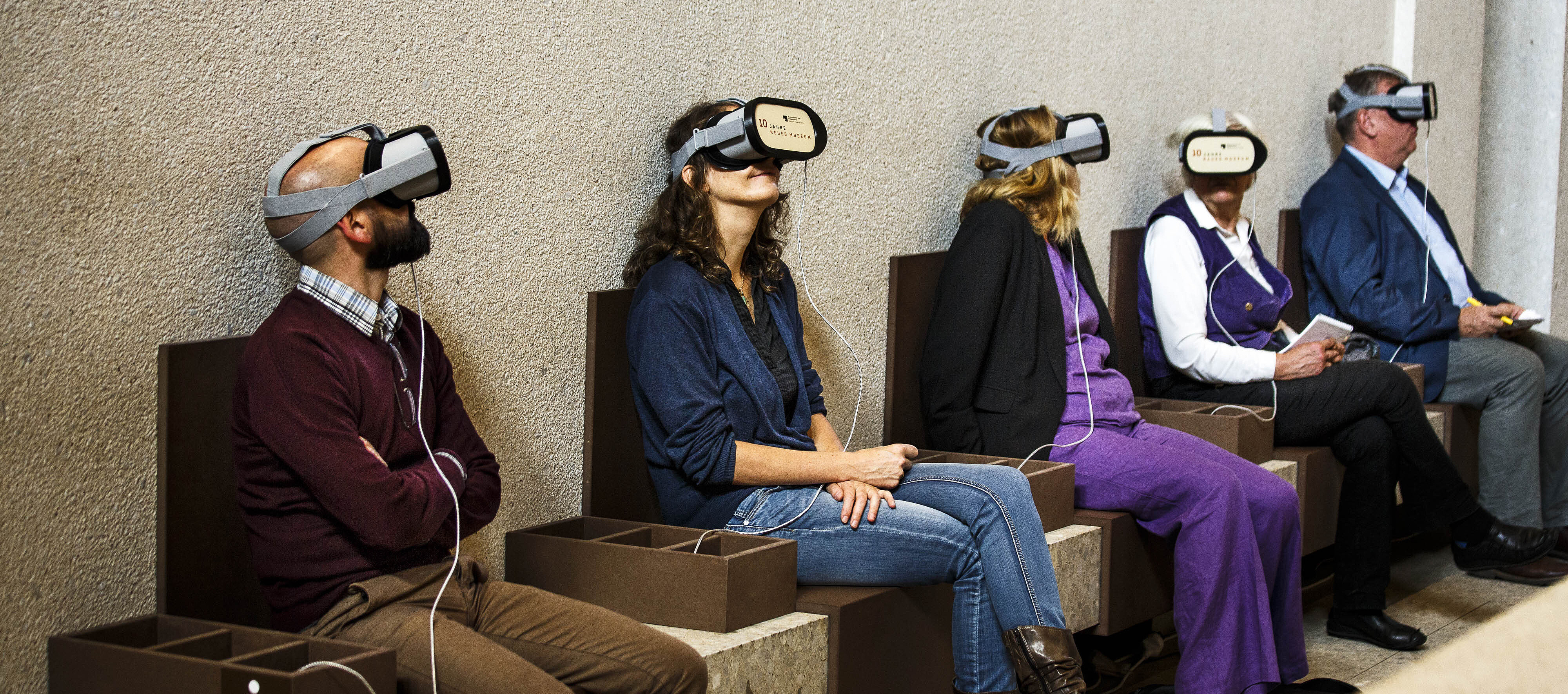 Fünf Personen unterschiedlichen Geschlechts und Alters mit Virtual-Reality-Headsets sitzen nebeneinander auf einer Bank in einem Raum