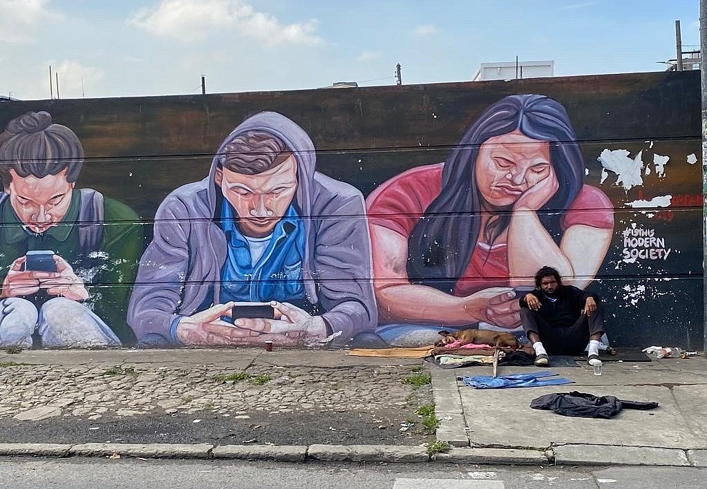 Wahrscheinlich obdachloser Mensch vor Graffiti-Wand mit Handynutzer_innen und Spruch "Is this modern society"