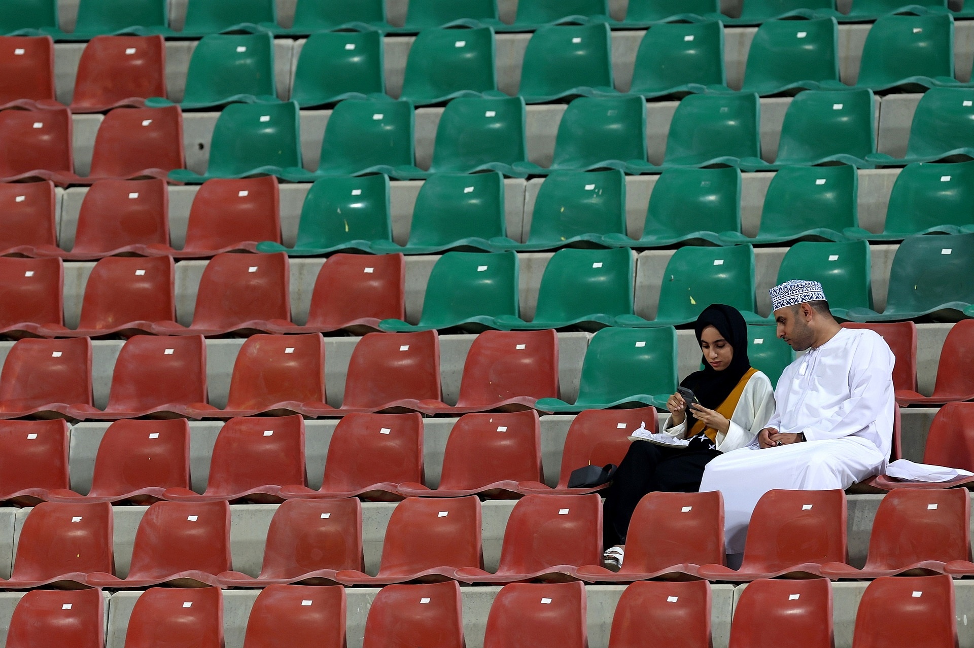 Mann und Frau sitzen in einem Fußballstadion im Oman. Alle anderen Plätze sind leer.