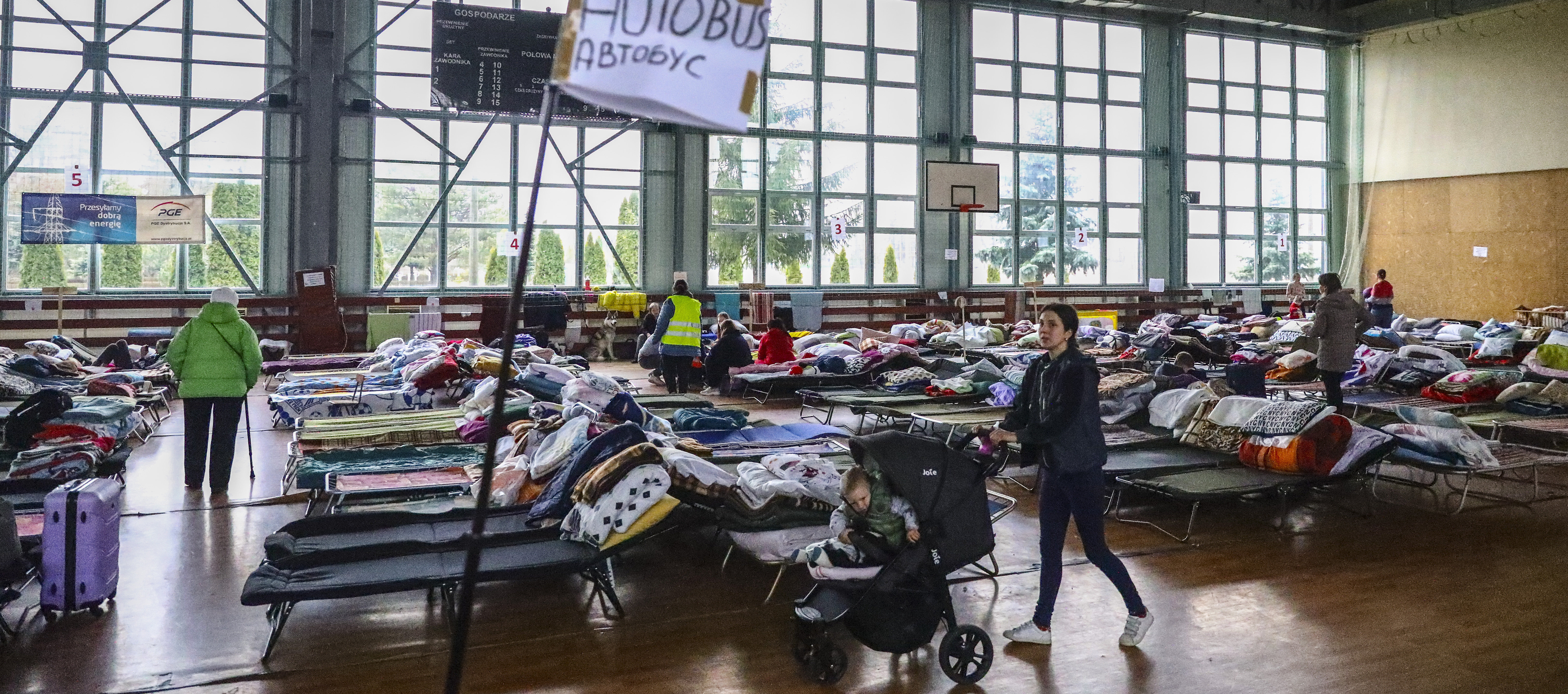 Flüchtlinge aus der Ukraine in einer Sporthalle in Polen. Feldbetten und ein Schild "Autobus" und Kinderwägen sind zu sehen.