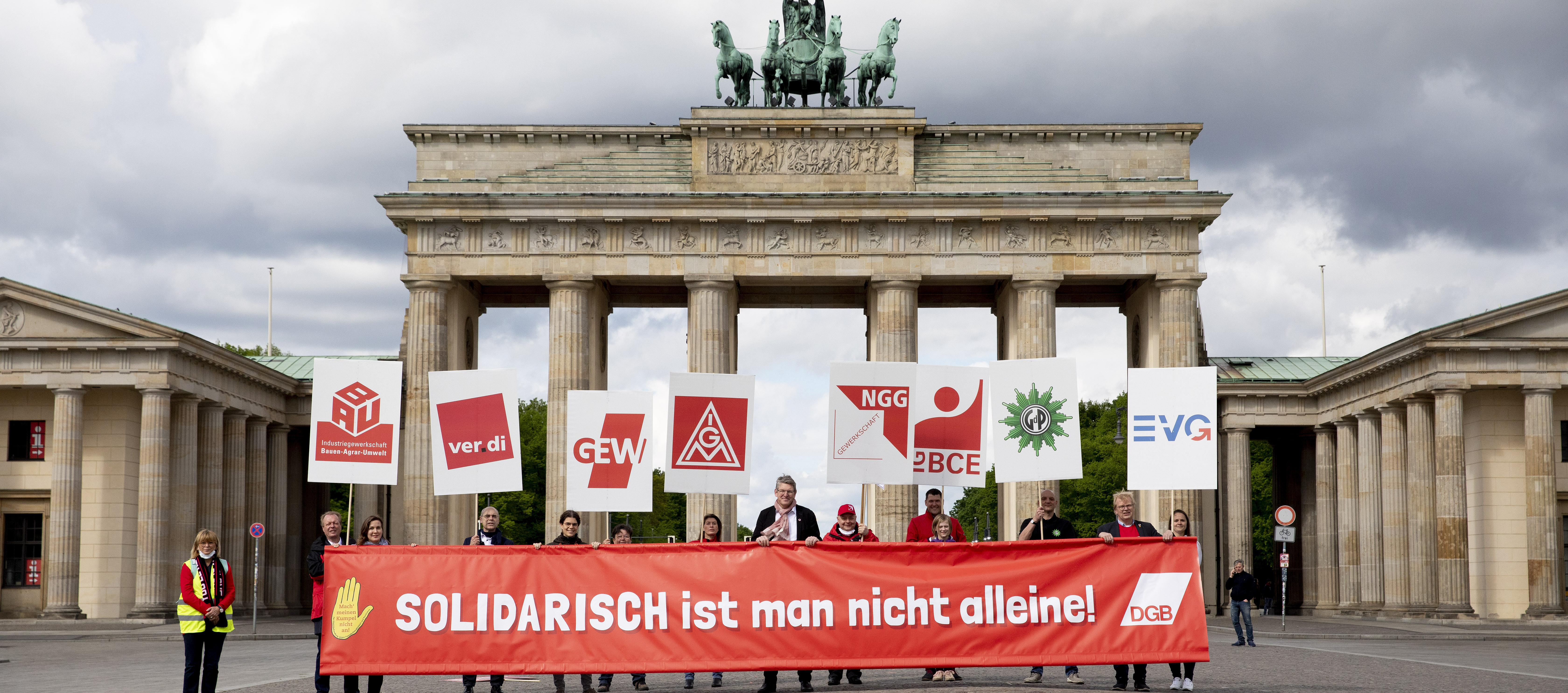 Foto mit Personen vor dem Brandenburger Tor. Zusammen halten Sie ein Transparent mit dem Schriftzug "Solidarisch ist man nicht alleine". Jede Person hält noch jeweils ein Plakat mit Gewerkschaftslogos.