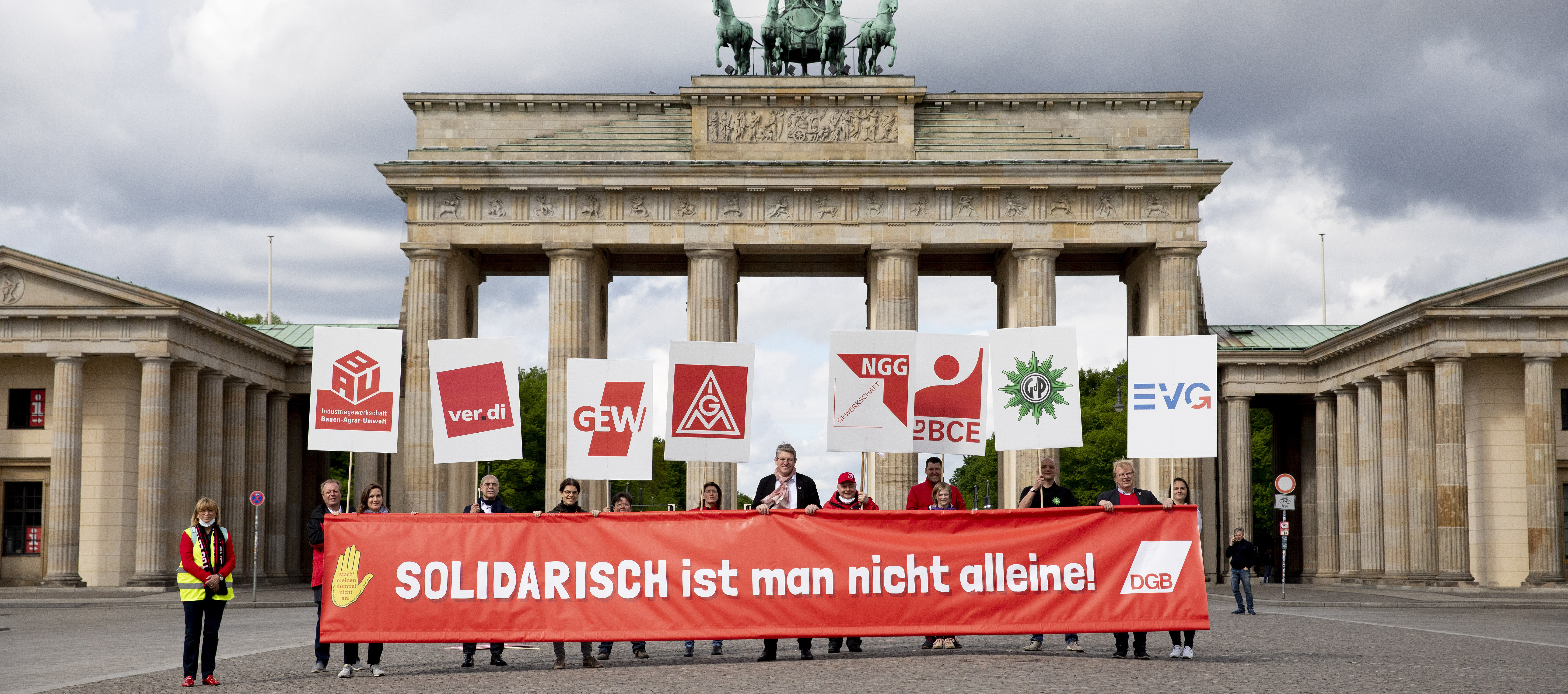 Foto mit Personen vor dem Brandenburger Tor. Zusammen halten Sie ein Transparent mit dem Schriftzug "Solidarisch ist man nicht alleine". Jede Person hält noch jeweils ein Plakat mit Gewerkschaftslogos.