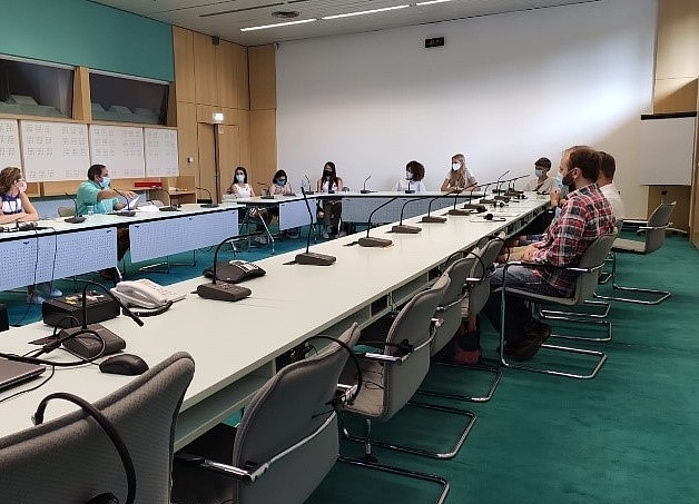 Teilnehmer:innen sitzen im Europarat in einem Besprechungsraum. Sie sitzten an Tischen im Kreis. Vor Ihnen sind Telefone und Mikros.