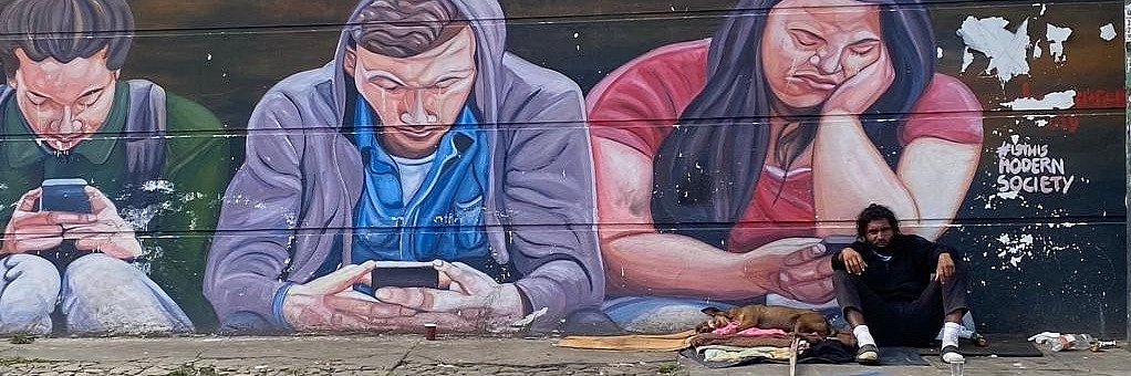 Wahrscheinlich obdachloser Mensch vor Graffiti-Wand mit Handynutzer_innen und Spruch "Is this modern society"