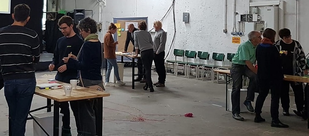 Das Bild zeigt Menschen um Tische herum stehend in einer Fabrikhalle. In der Halle ist außerdem eine Bühne aufgebaut.
