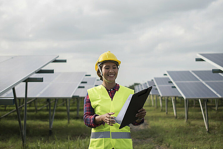 Lächelnde junge Frau mit Schutzhelm und Ordner zwischen Solaranlagen.
