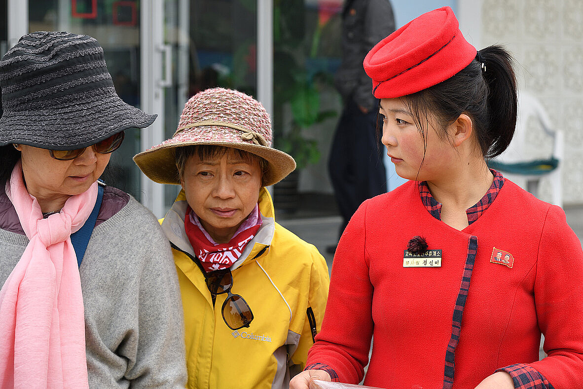 Zu sehen sind 3 ältere Frauen. Eine davon ist eine Frau in roter Uniform, die den beiden anderen etwas verkauft.