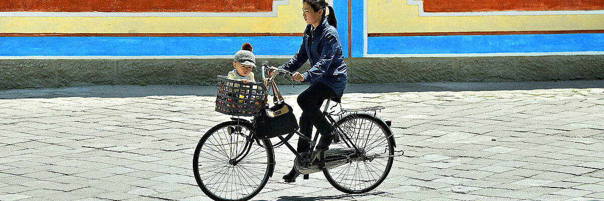 Im Vordergrund fährt eine Frau mit einem alten Fahrrad und einem Kind vorne im Einkaufskorb