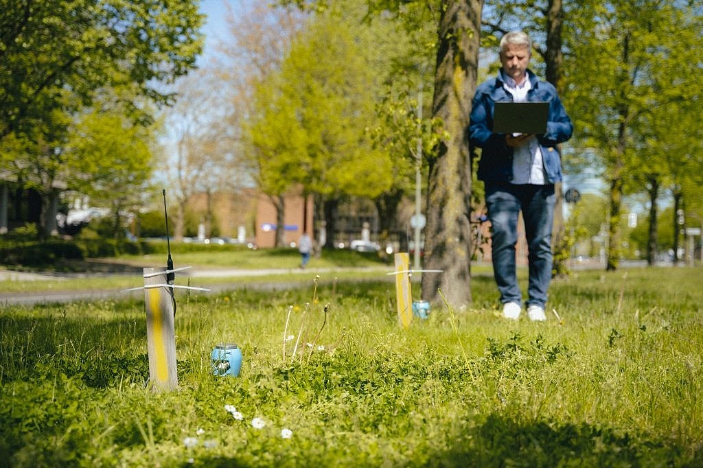 Bodenfeuchte-Messgerät auf einer Wiese in urbanem Umfeld. Im Hintergrund steht eine Person mit Laptop.