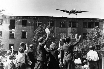 Schwarz-Weiß-Fotografie eines "Rosinenbombers" über winkenden Berlinern, 1948/1949. 