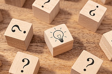 Symbolbild: Holzwürfel mit aufgedruckten Fragezeichen umkreisen einen Würfel mit dem Bild einer Glühbirne