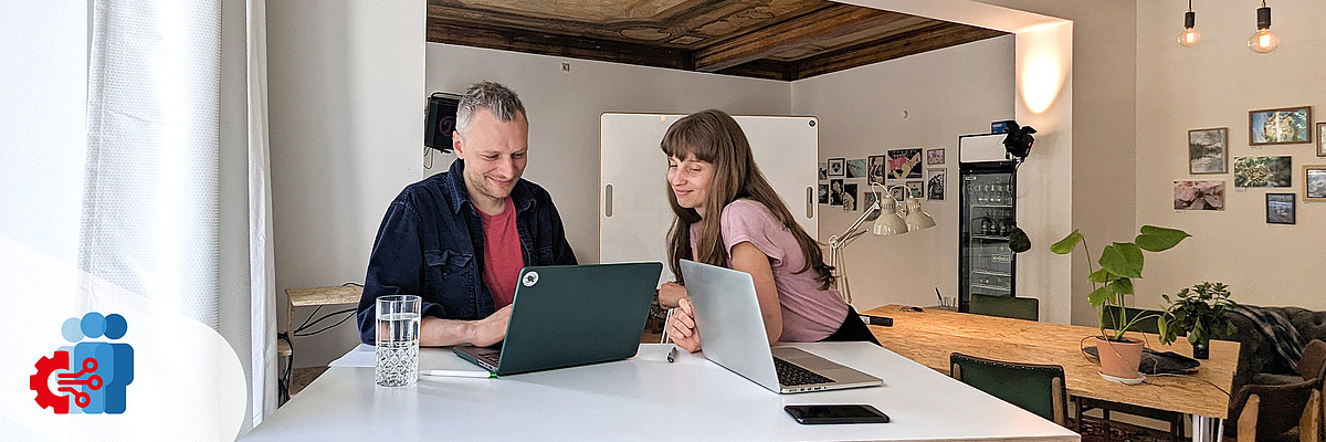 Ein Mann und eine Frau sitzen an einem weißen Tisch gemeinsam vor einem Laptop. Es handelt sich um einen Co-Working-Space mit modernem Mobiliar und Holzdecke.