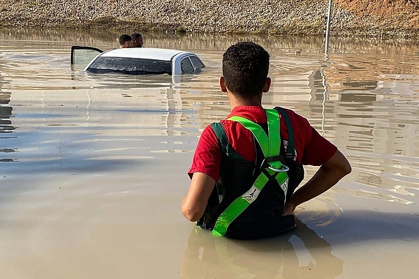 Rückenansicht eines Helfers der Organisation Libyscher Roter Halbmond, der bis zur Hüfte im Wasser steht und auf ein aus dem Wasser ragendes Autodach blickt 