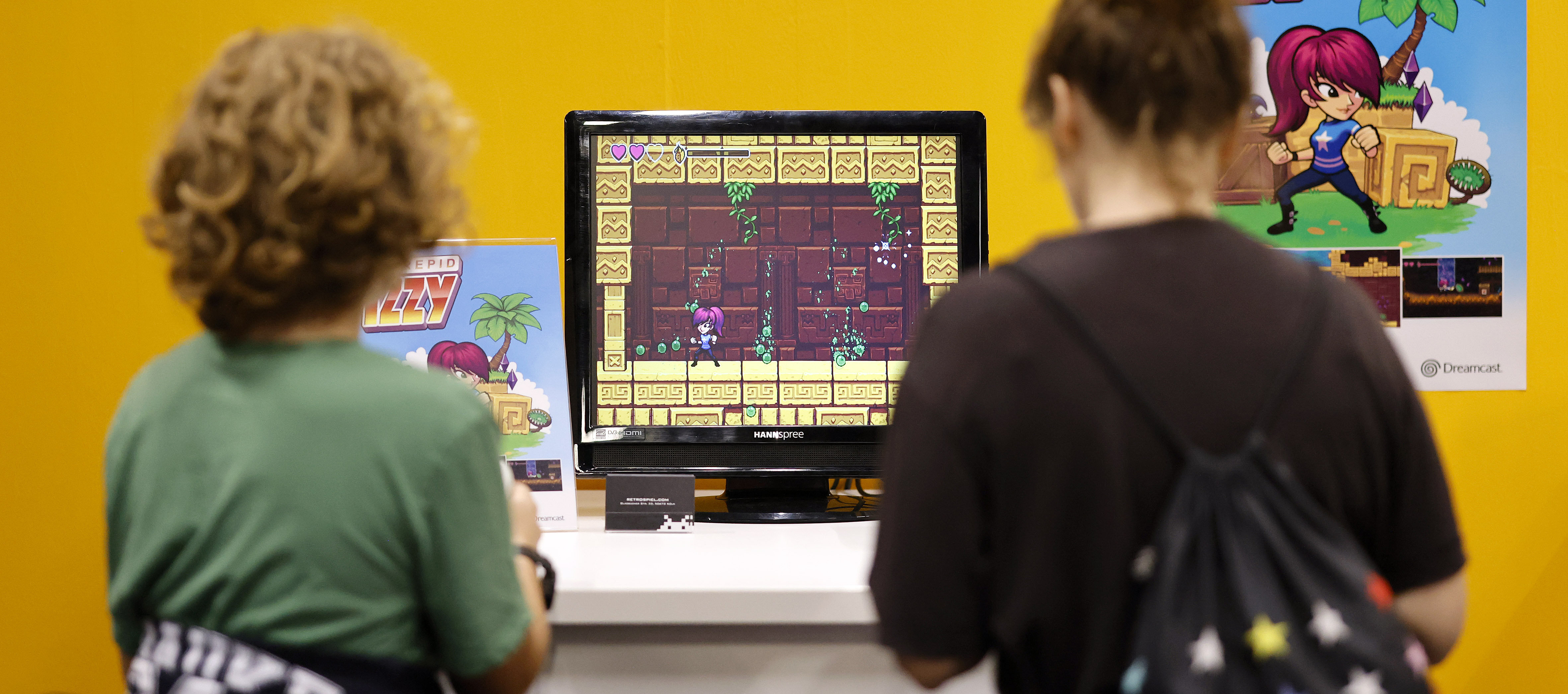 Zwei Jugendliche spielen ein Videospiel. Sie sind unscharf vor einem Schreibtisch vor einem orangefarbenen Hintergrund zu sehen. Zwei Plakate des Spiels sind zu erkennen, auf denen jeweils Protagonistin des Spiels (Eine weiblich gelesene Figure mit Pferdeschwanz) zu sehen ist. 