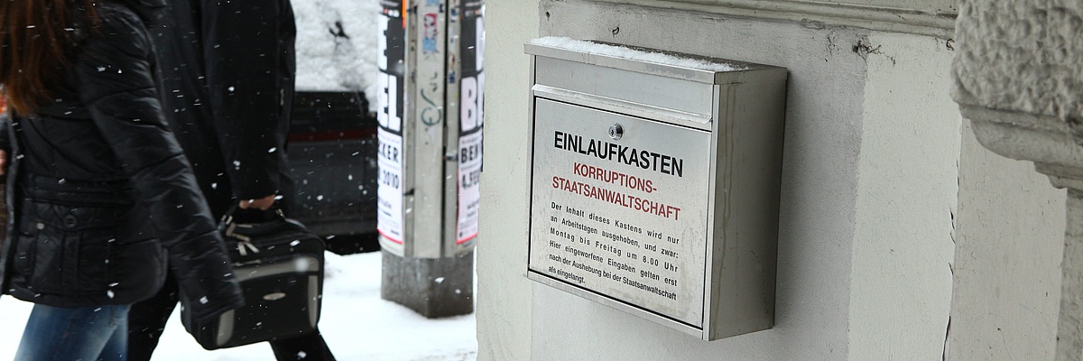 Hauseingang mit einem Briefkasten mit der Ausfschrift "Einlaufkasten - Korruptionsstaatsanwaltschaft". Im Hintergrund ein schneebedeckter Gehweg, auf dem zwei Personen vorbeigehen.