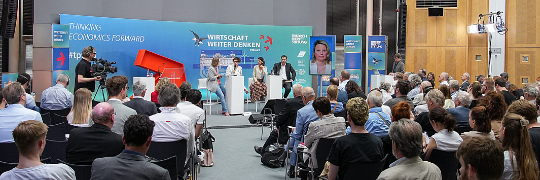 Bild einer Veranstaltung in der FES in Berlin. Zu sehen ist ein Publikum von hinten sowie eine Bühne mit mehreren Sprecher_innen und einer grün-blauen Rückwand.