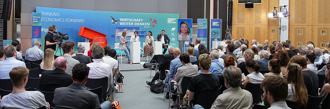 Bild einer Veranstaltung in der FES in Berlin. Zu sehen ist ein Publikum von hinten sowie eine Bühne mit mehreren Sprecher_innen und einer grün-blauen Rückwand.