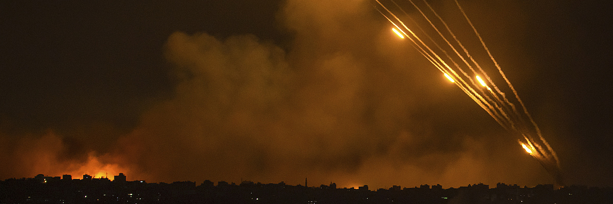 Mehrere Raketen, sichtbar als orange leuchtende Streifen am Nachthimmel, werden Richtung Israel gefeuert. Im Bildvordergrund bis zum Horizont ist eine Stadt im Dunkeln sichtbar.