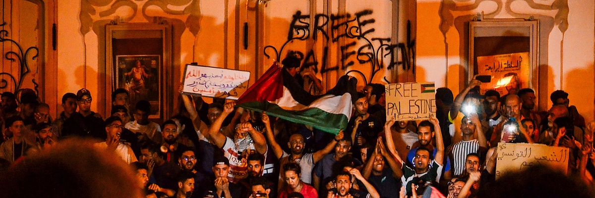 protestierende, vor allem junge Menschen vor einer Hauswand. Es wird eine palästinensiche Flagge hochgehalten und ein Schild mit der Aufschrift "Free Palestine"