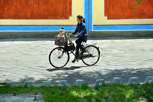 Im Vordergrund fährt eine Frau mit einem alten Fahrrad und einem Kind vorne im Einkaufskorb