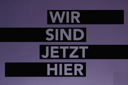 Text "Wir sind jetzt hier" weiß auf schwarzen Balken vor violettem Hintergrund