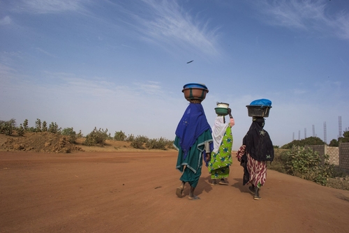 Afrikanische Frauen in traditioneller Kleidung gehen eine Straße entlang und tragen Töpfe auf dem Kopf.